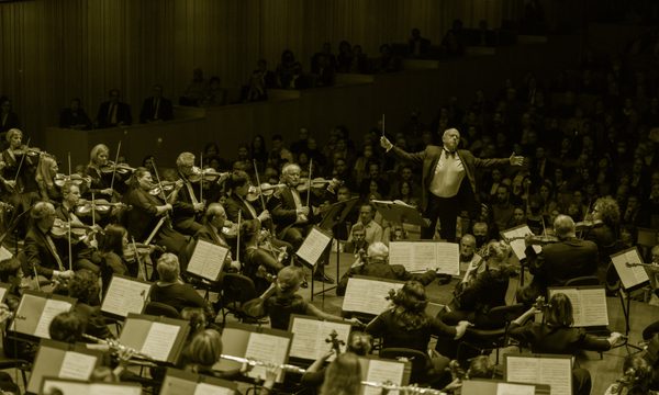 A conductor stands with his arms outstretched in the midst of an orchestra