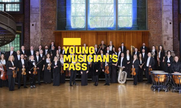 A yellow logo reading Young Musician's Pass overlays the orchestra standing posed with their instruments in a large room with big windows.