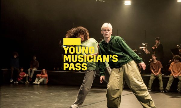 A yellow logo reading Young Musician's Pass overlays two dancers at the front of the stage, one is standing behind the other with their hand on the other dancer's shoulder. Behind them are other dancers seated and a group of musicians