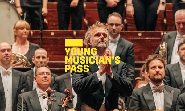 a A yellow logo reading Young Musician's Pass overlays a conductor standing on the platform facing the audience his hands together in gratitude
