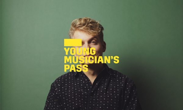 A yellow logo reading Young Musician's Pass overlays a man sitting facing the camera with one leg crossed over the other. He wears a black spotted shirt and jeans in front of a green background.
