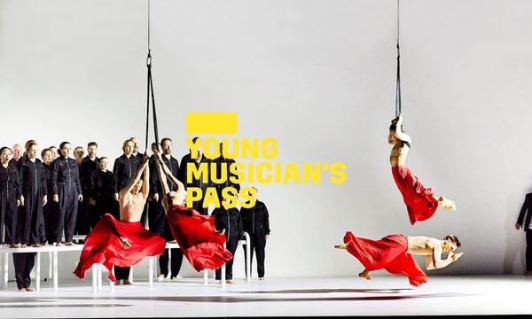 A yellow logo reading Young Musician's Pass overlays four dancers performing in red flowing skirts, three of which are suspended from ropes. In the background, a choir dressed in black jumpsuits sing on top of white platforms.