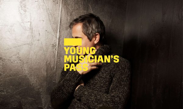 A yellow logo reading Young Musician's Pass overlays a man standing in a black room facing the side. He is wearing a knitted jumper and black trousers.