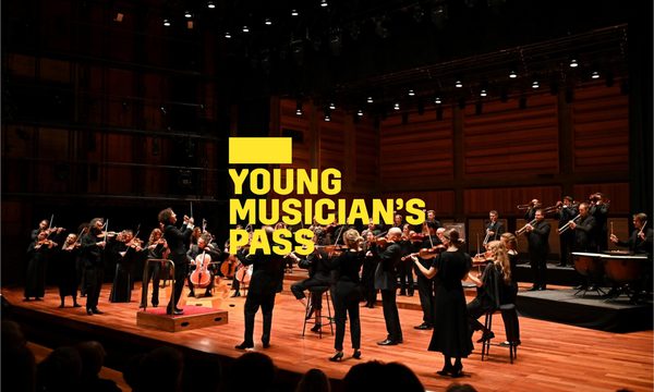 A yellow logo reading Young Musician's Pass overlays an orchestra dressed in black standing on stage playing their instruments, a conductor stands before them as an audience looks on
