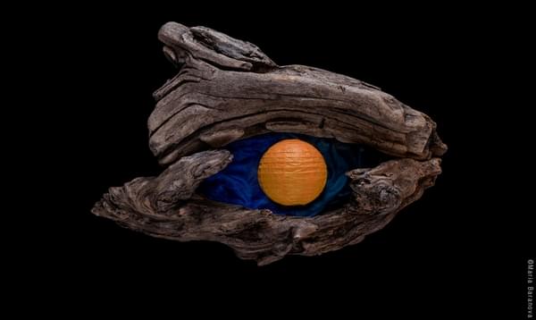 Driftwood encloses a spherical orange paper lantern lying on top of blue cloth. The arrangement resembles an eye.