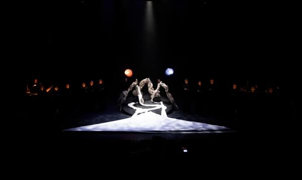 A triangle of white cloth lies in front of three arches made from gnarled wood. A blue and an orange paper lantern are held either side of the structure. The faces of choir members are illuminated in the darkness.