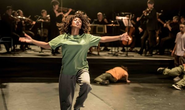 A dancer in a green t-shirt with her arms outspread. An orchestra can be seen behind her.
