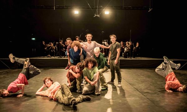 Dancers in casual clothes pose as a group, smiling, as two of their members breakdance either side.