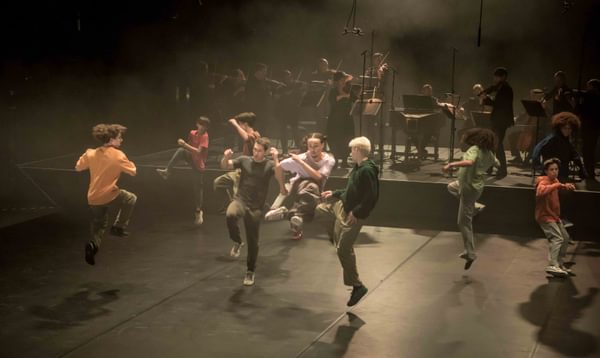 Dancers in casual clothing jump and kick their legs on stage.