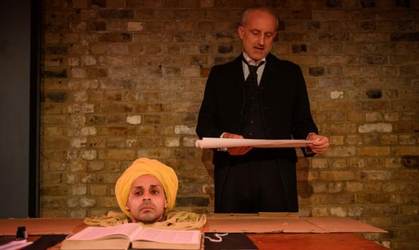 The disembodied head of a man in a yellow turban sits on a table in front of a book. Behind, a man in a black suit and tie reads from a scroll.