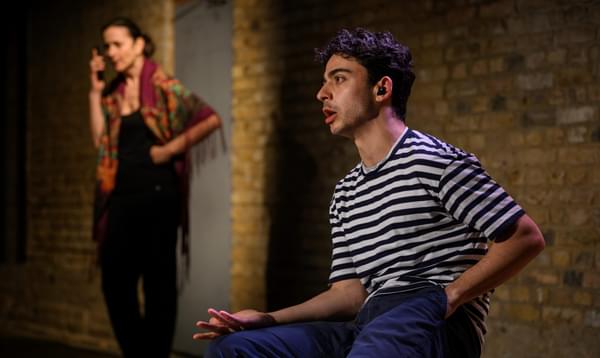 A man in a white stripy top sits and holds a palm up while a woman in a shawl takes a phone call in the background.