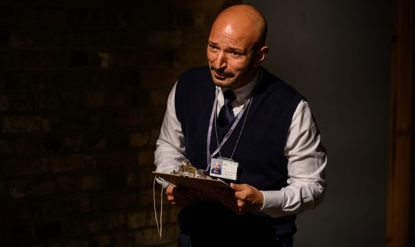 A bald man with a clipboard stood in the spotlight. He is wearing an ID badge.