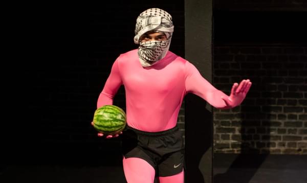 A person wearing a pink bodysuit and a keffiyeh holds up a hand while holding a watermelon.