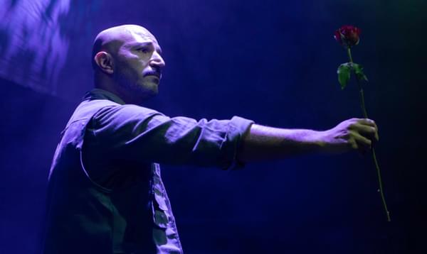 A sad man holds out a rose. Purple light shines on him.