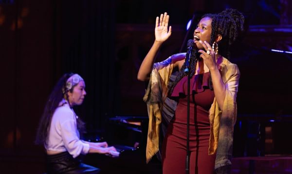 A woman sings and looks up with her hands raised. Another woman plays the piano behind her.