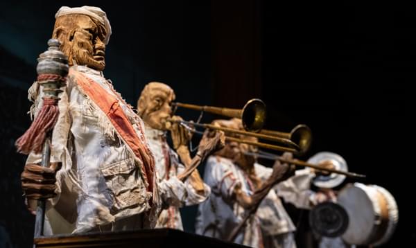 A puppet with a staff and a sash. Puppets playing trombones, cymbals, and drums stand behind the one holding the staff.