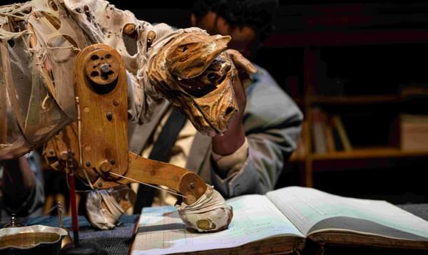 A hyena puppet made from wood and rags walks on top of a book, inspecting it.
