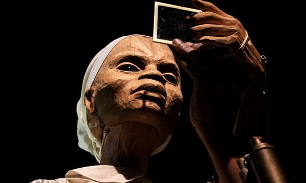 The puppet of a woman closely inspects a polaroid photo.