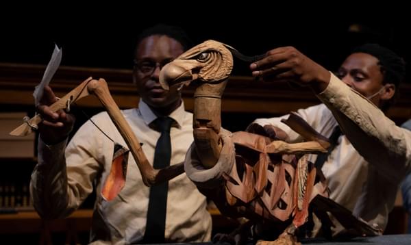 An angry looking wooden vulture puppet. Two puppeteers stand behind it, manipulating the puppet.