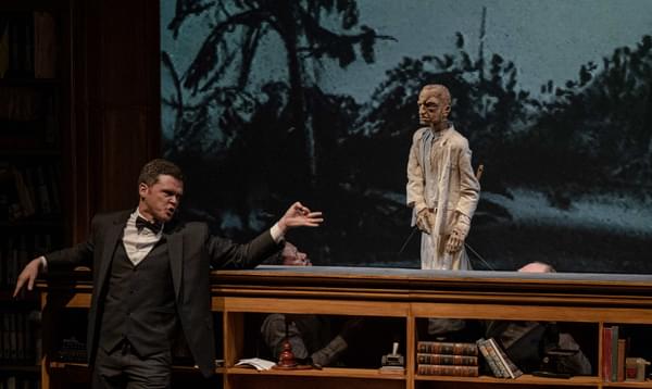 A man in a suit and bowtie gestures at a puppet dressed in white clothing.