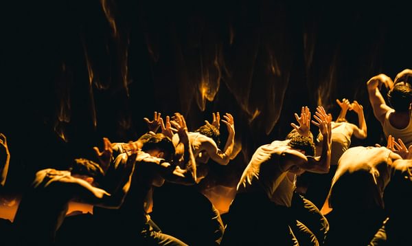 Performers lit by gold light bow their heads and hold their hands up