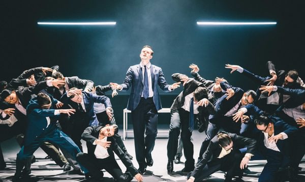 A man in a suit stands in the spotlight, as many other performers in business attire pose either side of him
