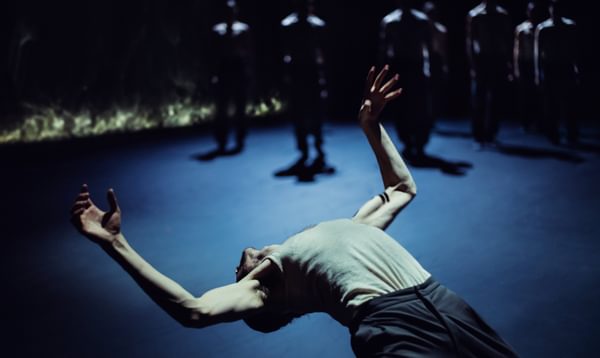 A dancer leans back far enough that their head is upside down and their front torso arched. Their arms flex above them. Other performers are silhouetted in the background.
