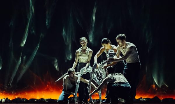 Five performers in vests puppet the skeleton of a large dog. Their backdrop resembles smoking coals.
