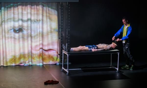 A man wearing shorts lies on a table while another man points small camera directly at his face. A projection of the lying man's face is shown on a curtain.