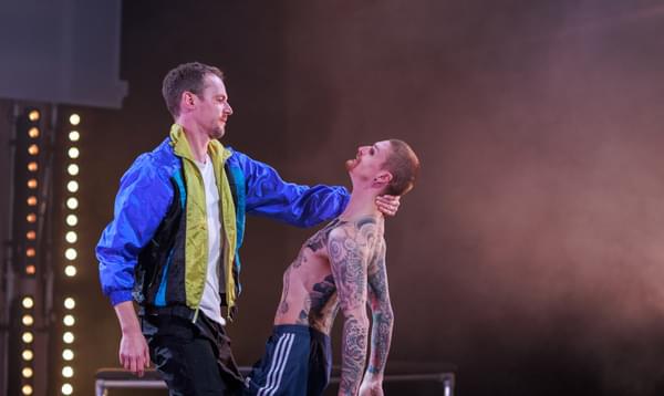 A man in a blue jacket holds another man by the back of his neck so that they face each other. The man being held is shirtless and covered in tattoos.