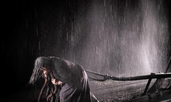A woman in a shawl drags a plough using thick ropes, while rain pours down.