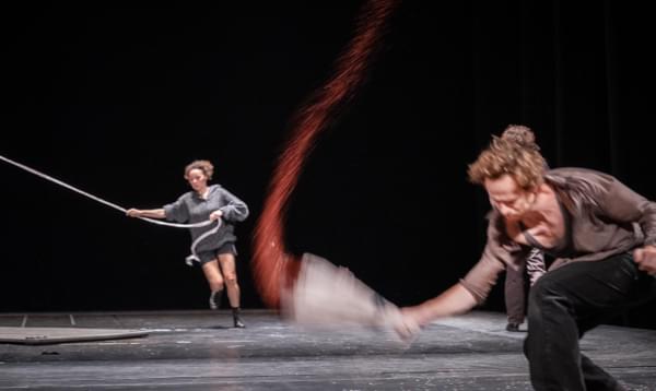 A man swings something at the ground and a trail of red dust arcs through the air. In the background a woman runs with a rope held taut.
