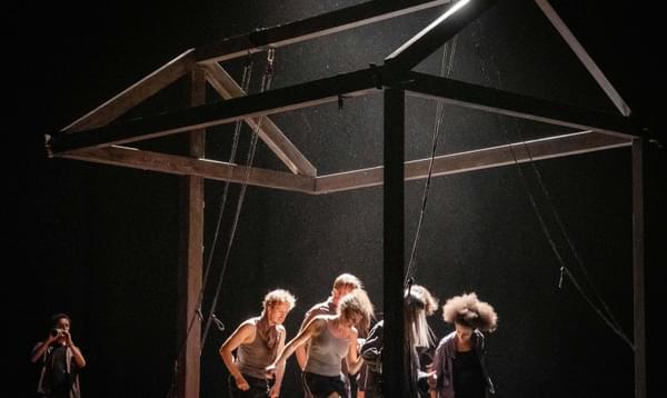 A group of performers dance and move within the frame of a house.