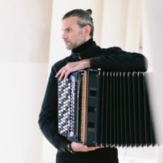 A serious looking man faces left holding a black accordion.