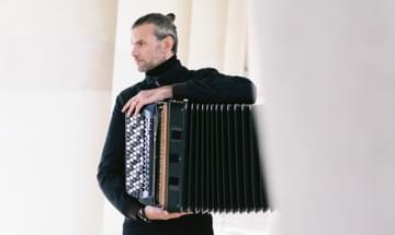 A serious looking man faces left holding a black accordion.