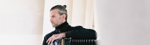 A serious looking man faces left holding a black accordion.