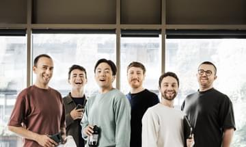 Six singers stand facing the camera dressed in casual clothes
