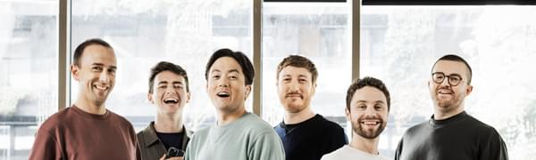 Six singers stand facing the camera dressed in casual clothes
