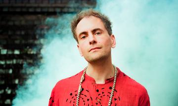 A man seen from the shoulders up looking at the camera with a neutral expression, standing in front of a pale blue cloud.