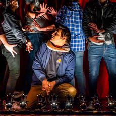 A man crouches on stage looking up at a group of people standing behind him, they are reaching their hands out towards him