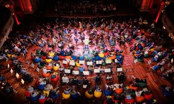 A birdseye view of an audience sitting on colourful beanbags with musicians placed among them and the conductor standing in the centre
