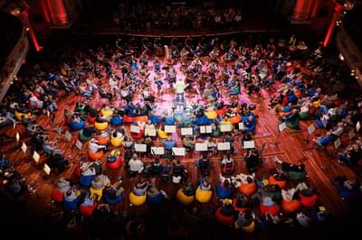 A birdseye view of an audience sitting on colourful beanbags with musicians placed among them and the conductor standing in the centre