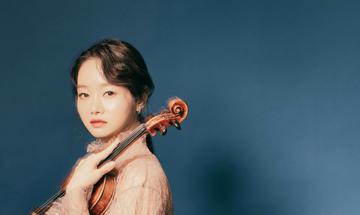 A woman looks at the camera facing side on, she is holding a violin on her shoulder and wearing a pink dress, while standing in front of a blue background