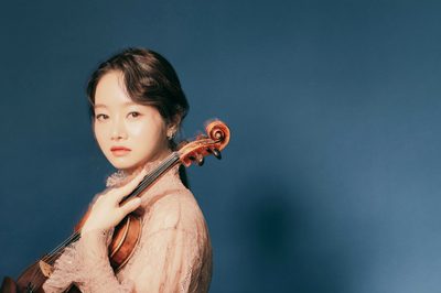 A woman looks at the camera facing side on, she is holding a violin on her shoulder and wearing a pink dress, while standing in front of a blue background