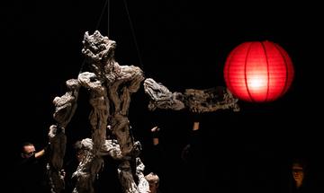 A large puppet with a rough rocky texture is being held up by 4 people. A red paper lantern hangs in front of them.
