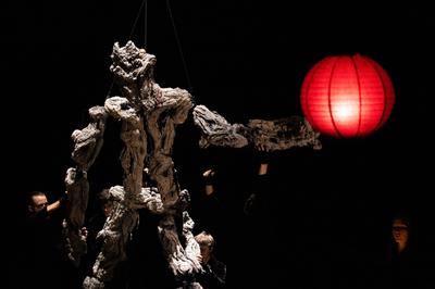 A large puppet with a rough rocky texture is being held up by 4 people. A red paper lantern hangs in front of them.