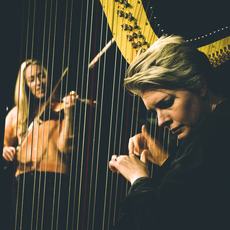 A woman sits facing the side while playing the harp. Behind her is a woman standing playing the violin
