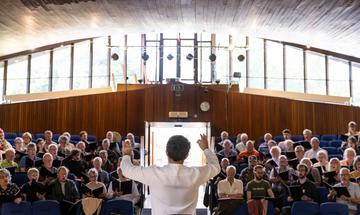 A conductor stands before a chorus with his back to the camera with his arms raised