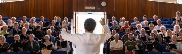 A conductor stands before a chorus with his back to the camera with his arms raised