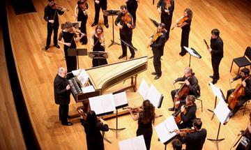 An ensemble of musicians dressed in black stand on stage playing string instruments. The conductor stands in front of them beside a piano.